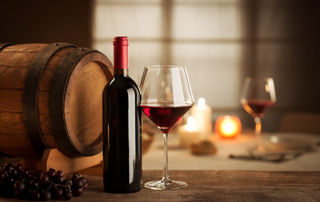 Ein Glas gefüllt mit Rotwein, sowie Flasche und Trauben auf einem Tisch. Daneben ein Barriquefass, das eine Rolle für die Tannine in Weinen spielt.