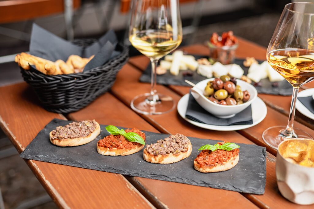 Vier Bruschetta-Scheiben mit verschiedenen Cremes im Vordergrund, außerdem zwei Gläser Wein sowie weitere Antipasti dahinter