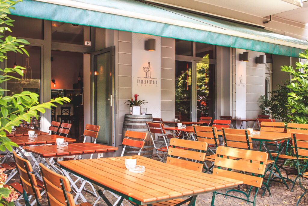 Außenansicht der Barrel Winebar, eine Terrasse überdacht von einer Markise, auf der viele Tische und Stühle angeordnet sind