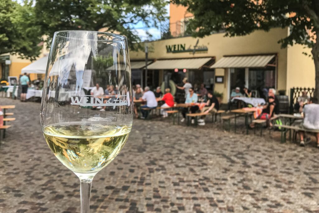 Im Vordergrund ein Weinglas mit dem Logo des Lokals "Wein & fein". Im Hintergrund sitzen mehrere Menschen auf der Terrasse und genießen ein Glas Wein bei schönem Wetter.