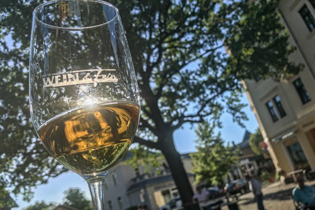 Die Sonne strahlt durch das Weinglas vor der Kamera, in dem sich Weißwein befindet.