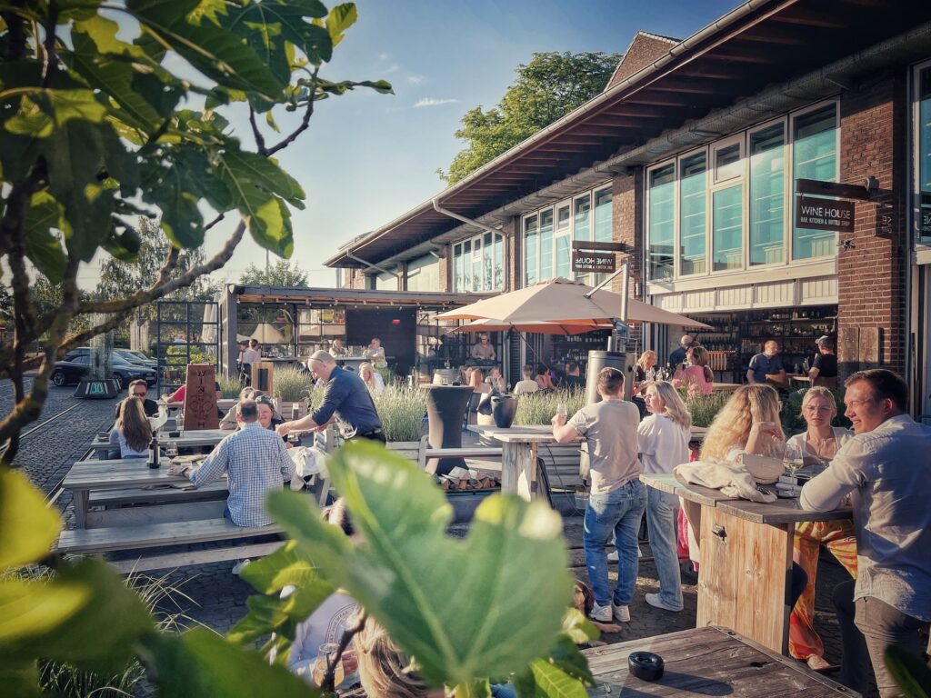 Die Terrasse des Wine House. Bei gutem Wetter sitzen und stehen viele Menschen an Holztischen und trinken ein Glas Wein