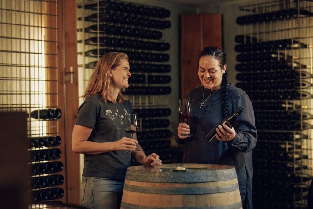 Die beiden Winzerinnen Meike und Dörte Näkel mit einem Glas Wein in der Hand in ihrem Weinkeller. Sie stehen an einem Fass und unterhalten sich