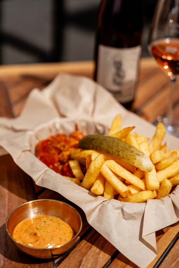 Eine Portion Pommes mit einer Essiggurke darauf. Daneben ein Glas Weißwein